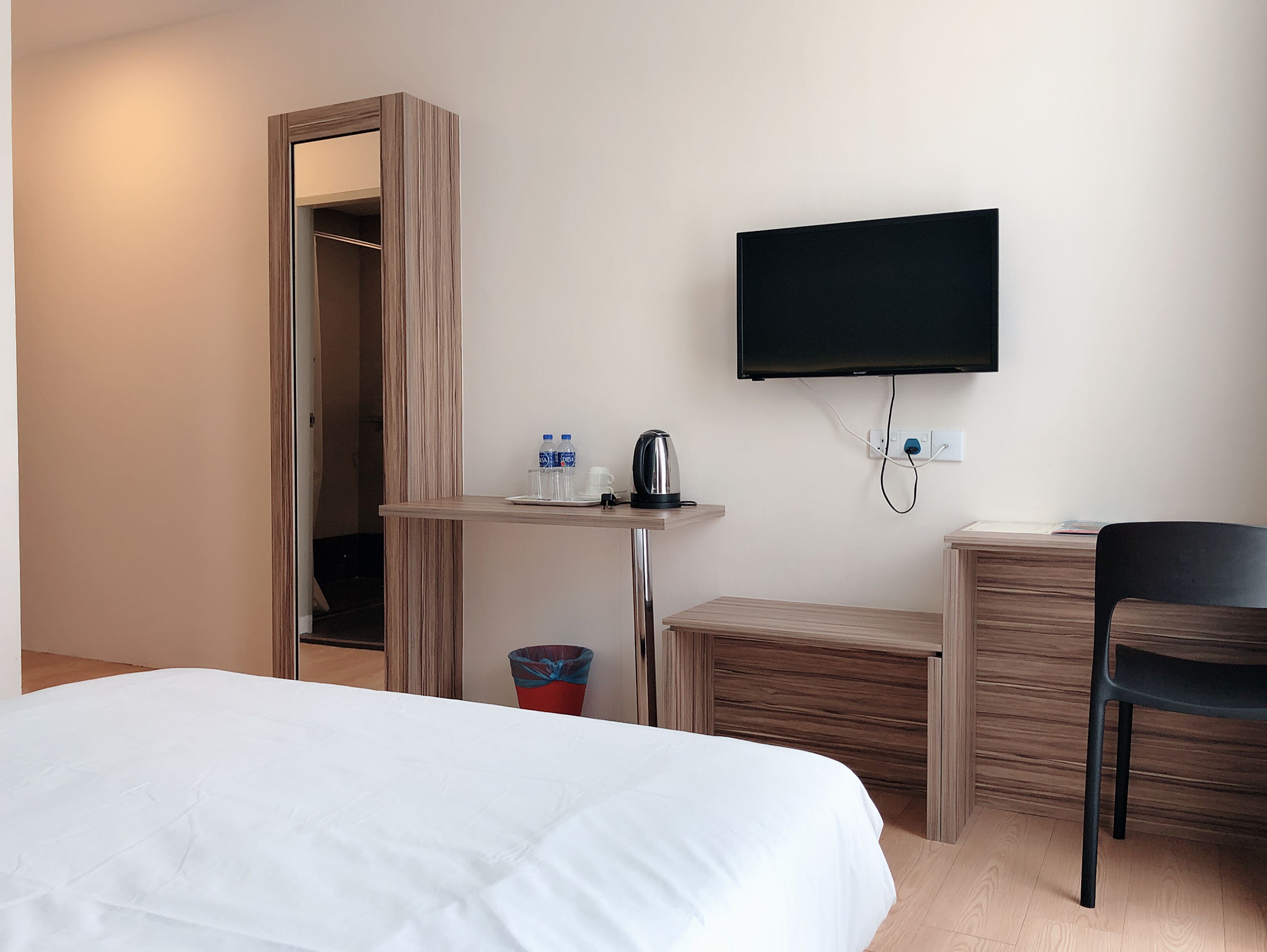 A hotel room with a bed, a desk with a flat-screen TV, a chair, and a small table with a kettle and water bottles.