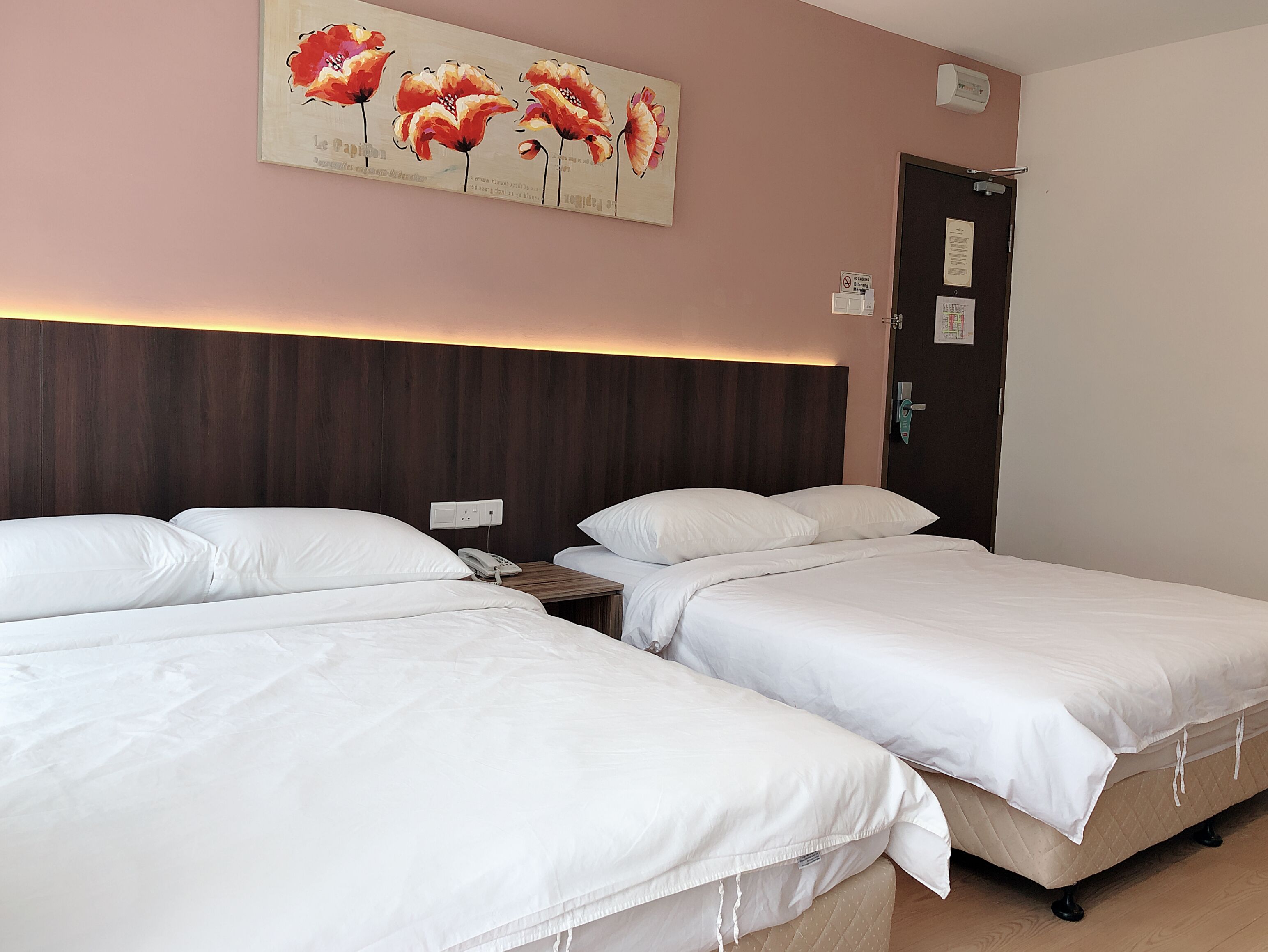 A hotel room with two beds, a wooden headboard, a painting of flowers, a phone, and a door with a sign.