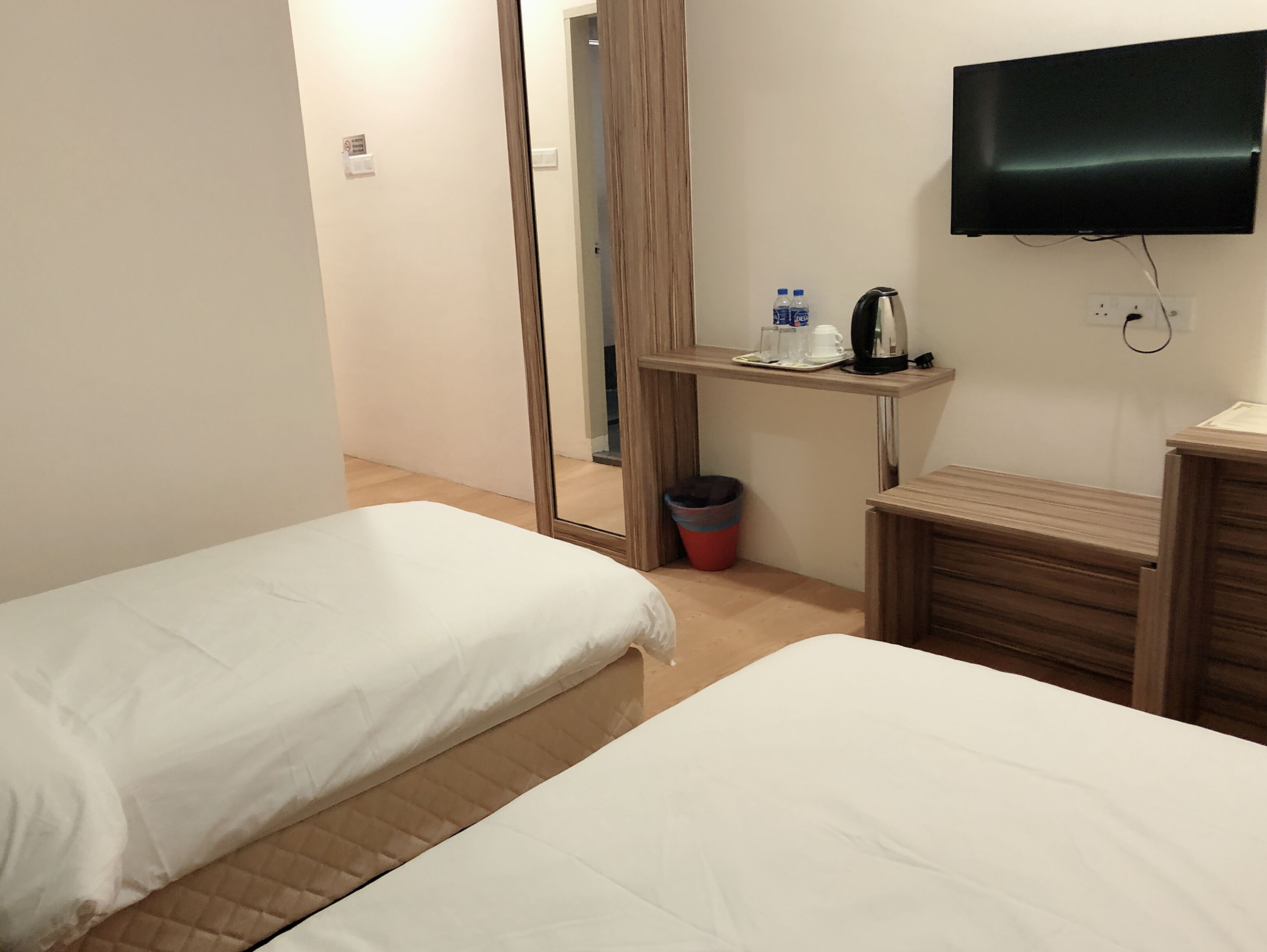 A hotel room with two beds, a TV, a small table with a kettle and cups, and a trash bin.