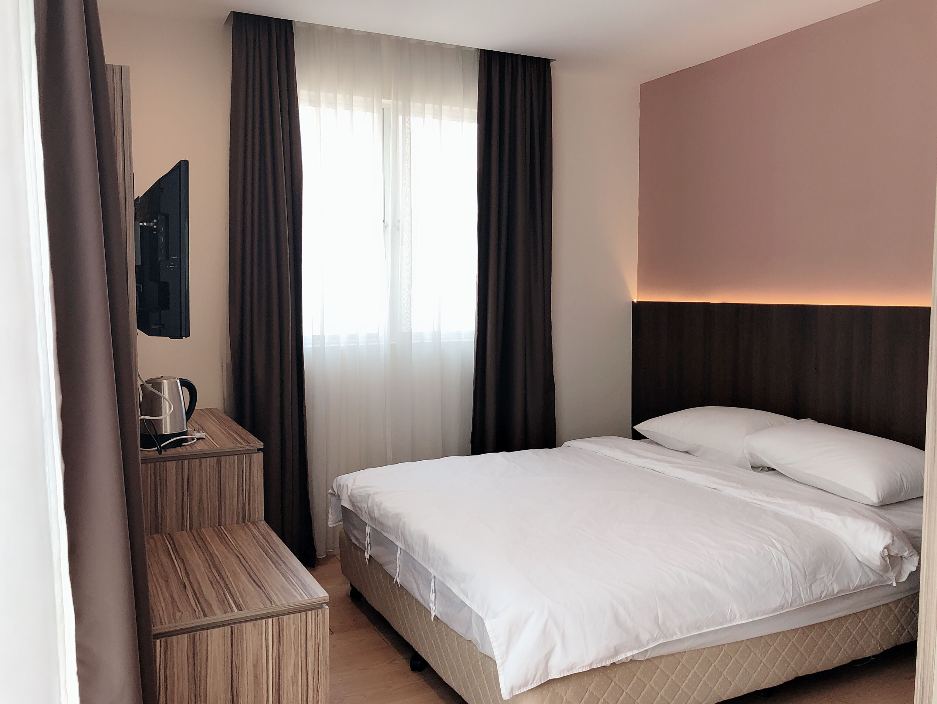 Ein Hotelzimmer mit Bett, Nachttisch aus Holz, Flachbildfernseher und einem Fenster mit Vorhängen.