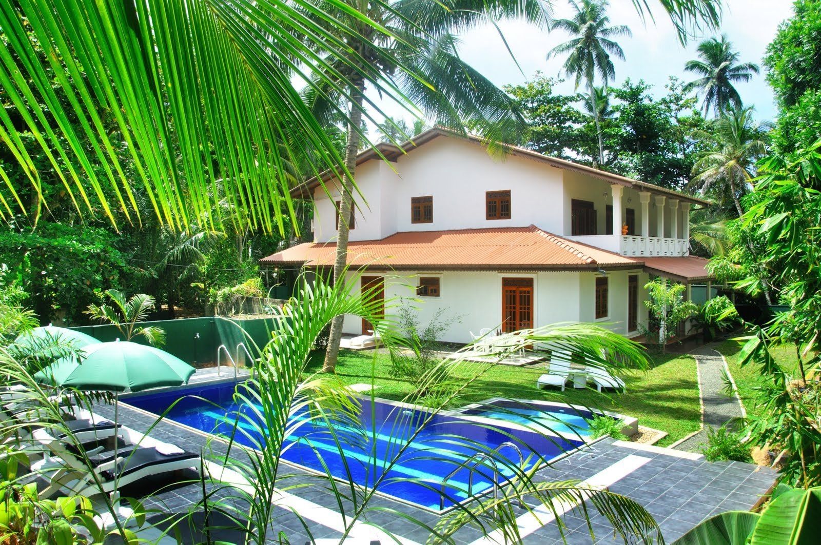 A two-story house with a swimming pool, surrounded by lush greenery and palm trees.
