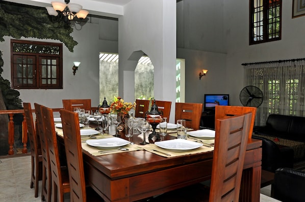A dining area with a wooden table set for six, surrounded by chairs, with a television and a fan in the background.