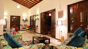 A spacious living area with a high wooden ceiling, a sofa with colorful cushions, a small table with decorative items, and a doorway leading to another room. - Hikka Resort (Hikkaduwa)