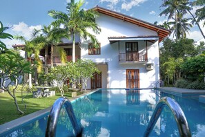 Una piscina con escalera metálica, rodeada de abundante vegetación y un edificio de dos pisos con balcones.
