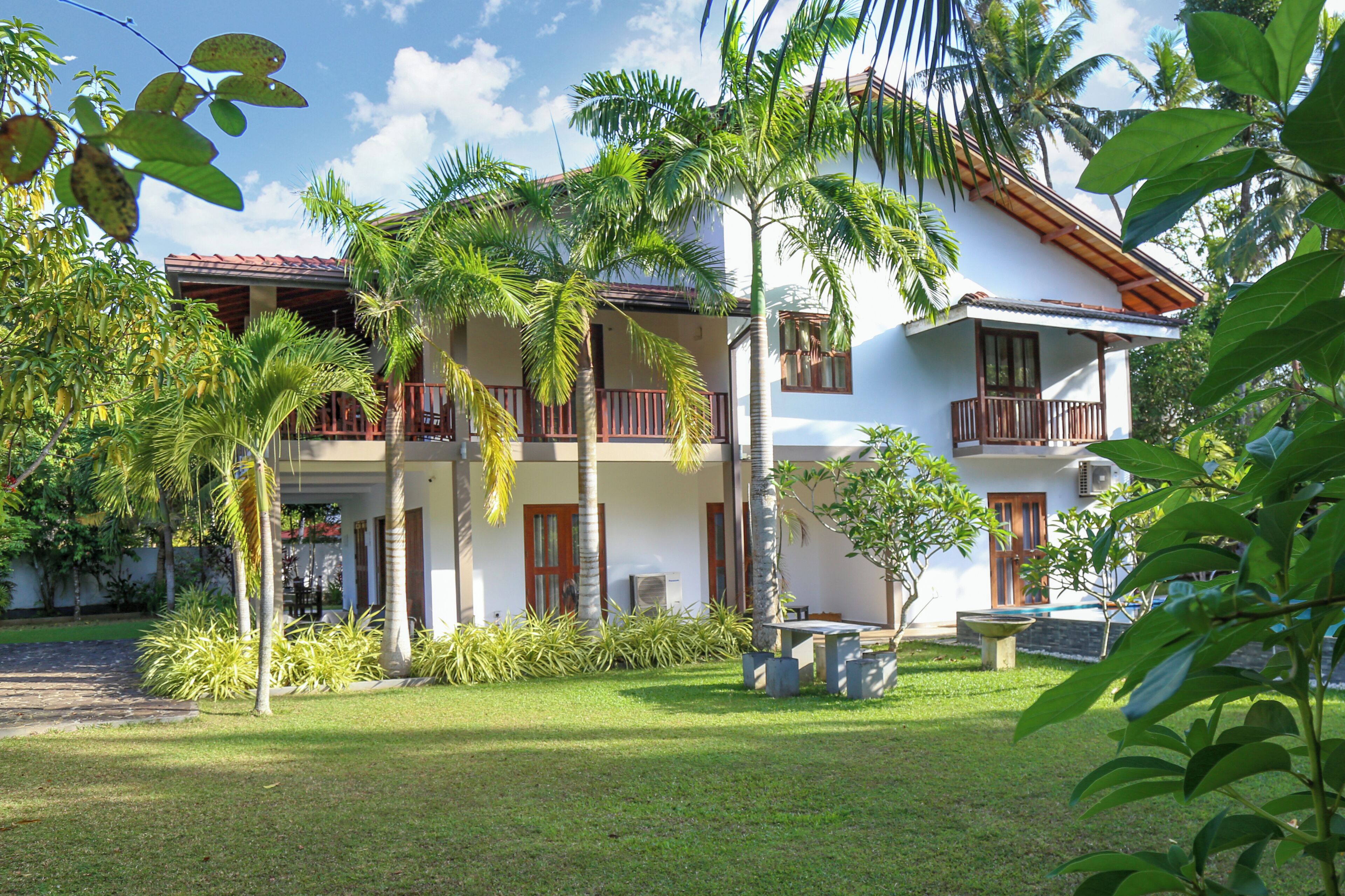A two-story building with a white facade, wooden balconies, and a well-maintained garden with palm trees and shrubs.