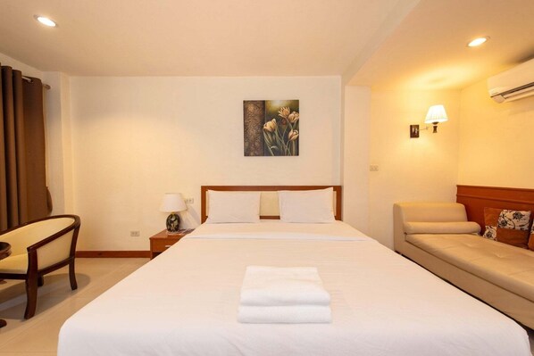 A hotel room with a large bed, a bedside table with a lamp, a chair, a sofa, and a painting on the wall. - Nuntiya Terrace (Udon Thani)