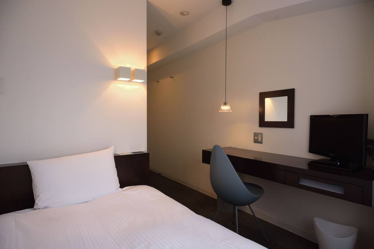 A hotel room with a bed, a desk with a chair, a television, and a wall-mounted mirror.