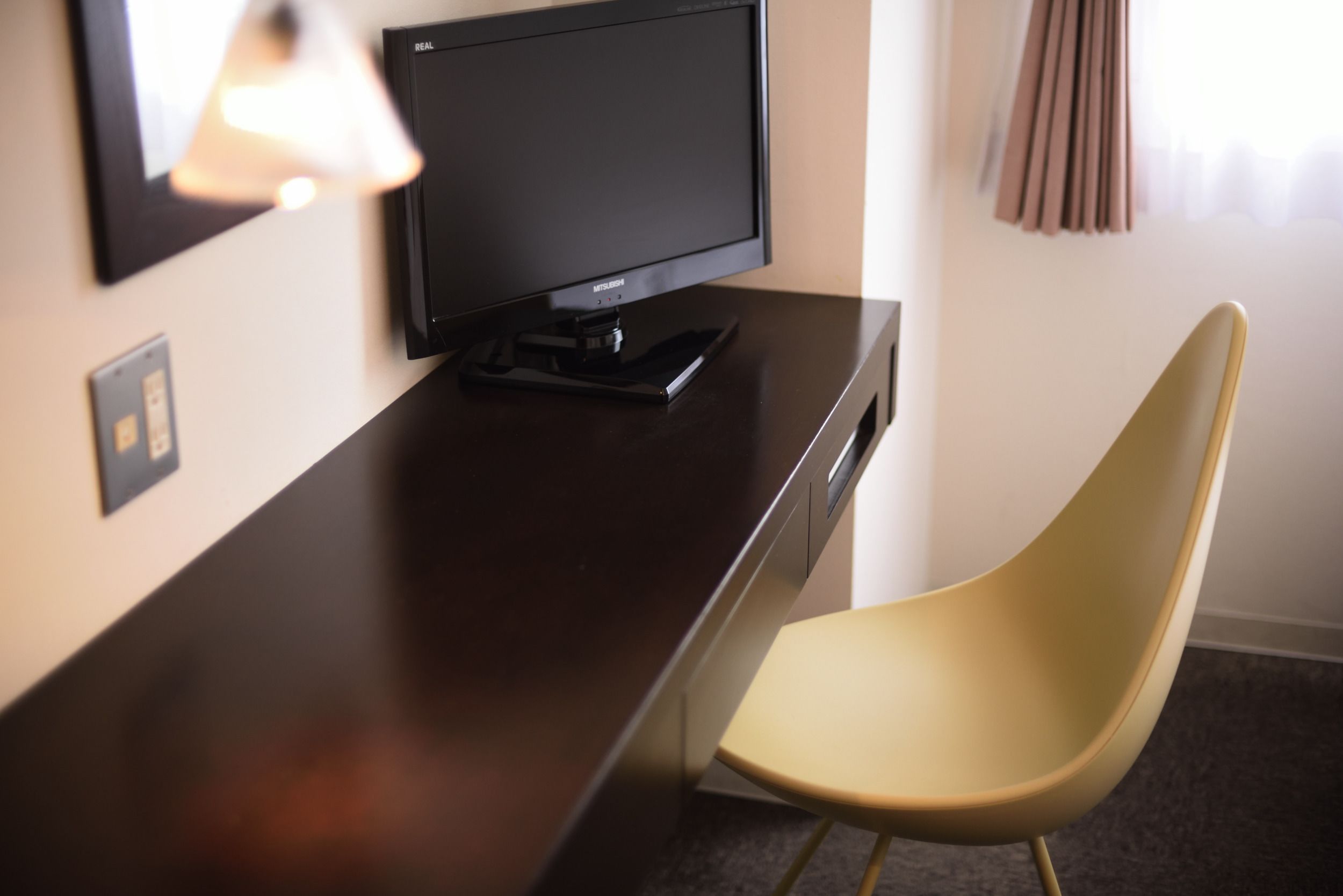 A modern hotel room with a dark wooden desk, a black flat-screen TV, a yellow chair, and a window with curtains.