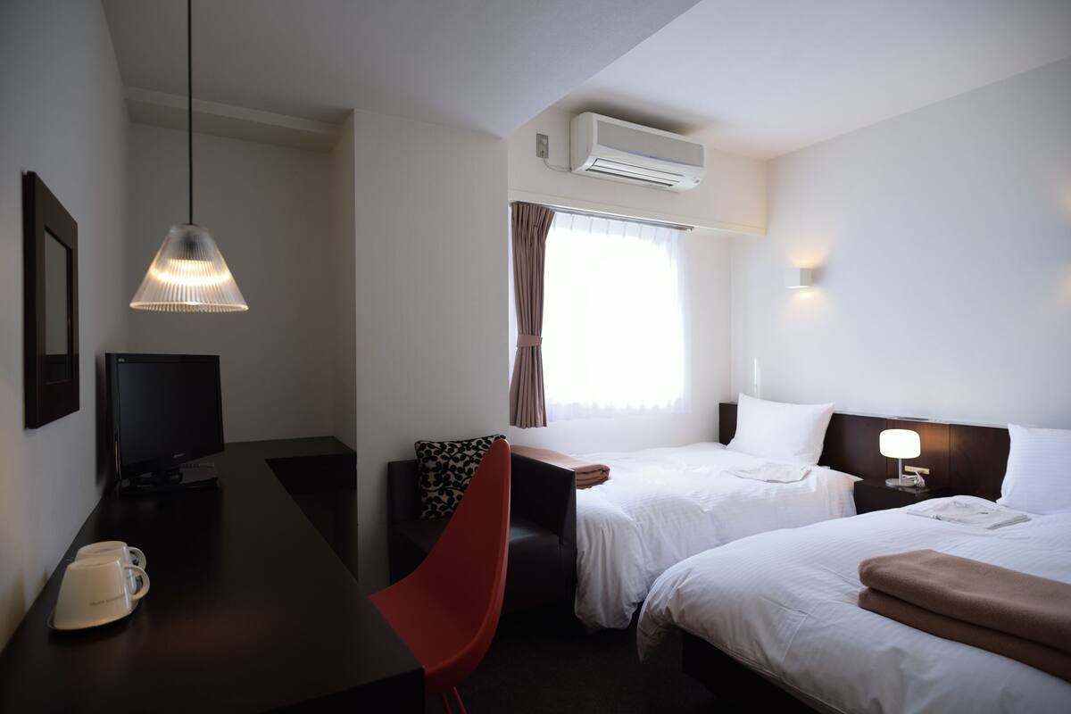 A hotel room with two beds, a desk with a computer, a red chair, a TV, and an air conditioning unit.