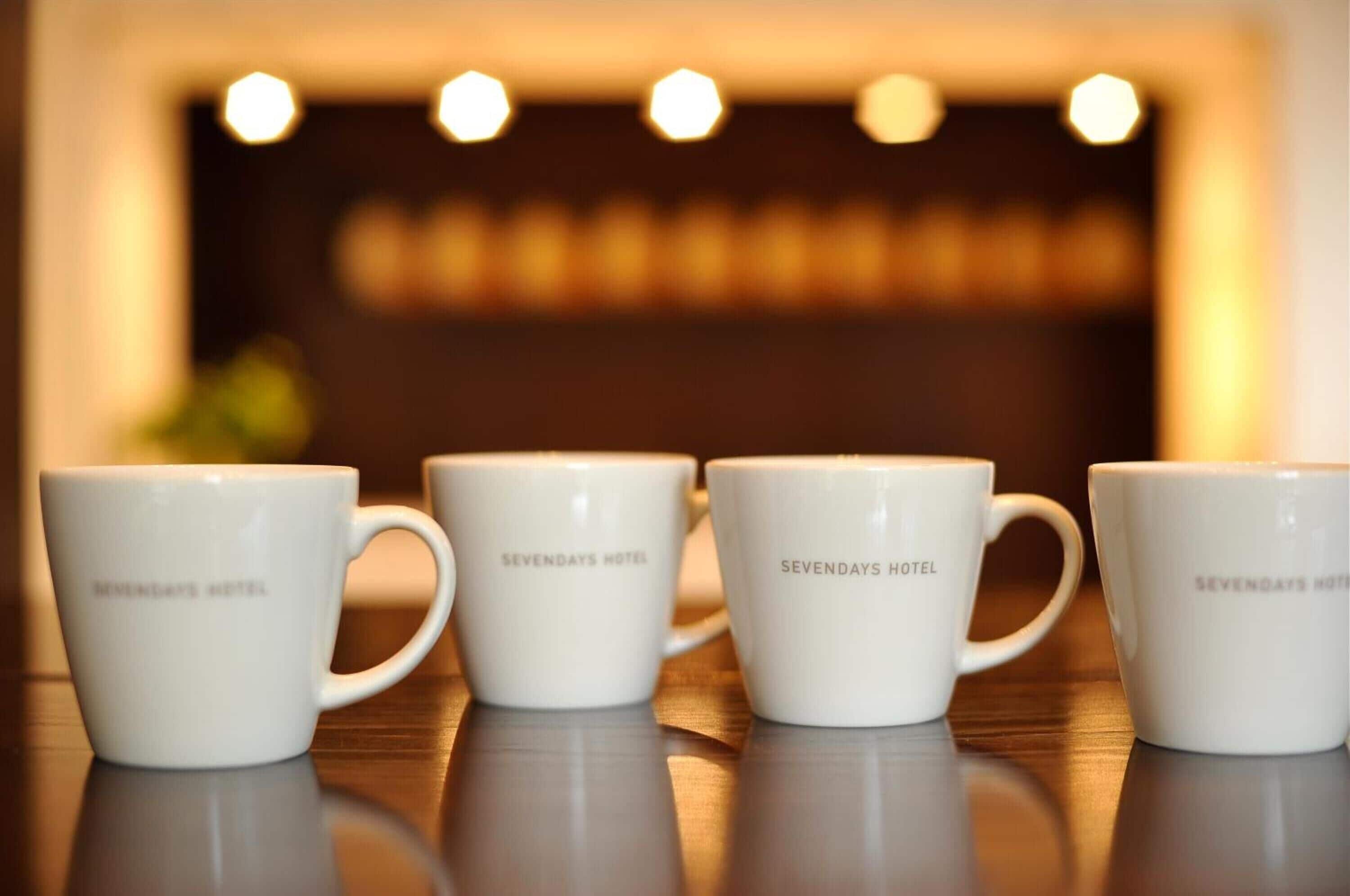 Four white ceramic mugs with "SEVEN DAYS HOTEL" text, arranged on a reflective surface.