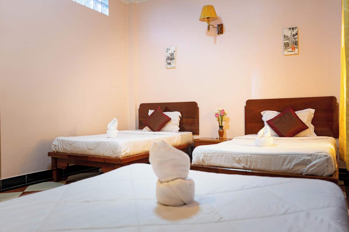 Une chambre d’hôtel avec deux lits simples, chacun doté d’une tête de lit en bois, de draps blancs et d’un oreiller rouge. Une table de chevet avec une lampe et un vase de fleurs se trouve entre les deux lits.