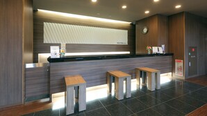 A modern reception area with a wooden reception desk, two wooden stools, a clock, and a fire alarm. - Hotel Route-Inn Shimizu Inter (Shizuoka)