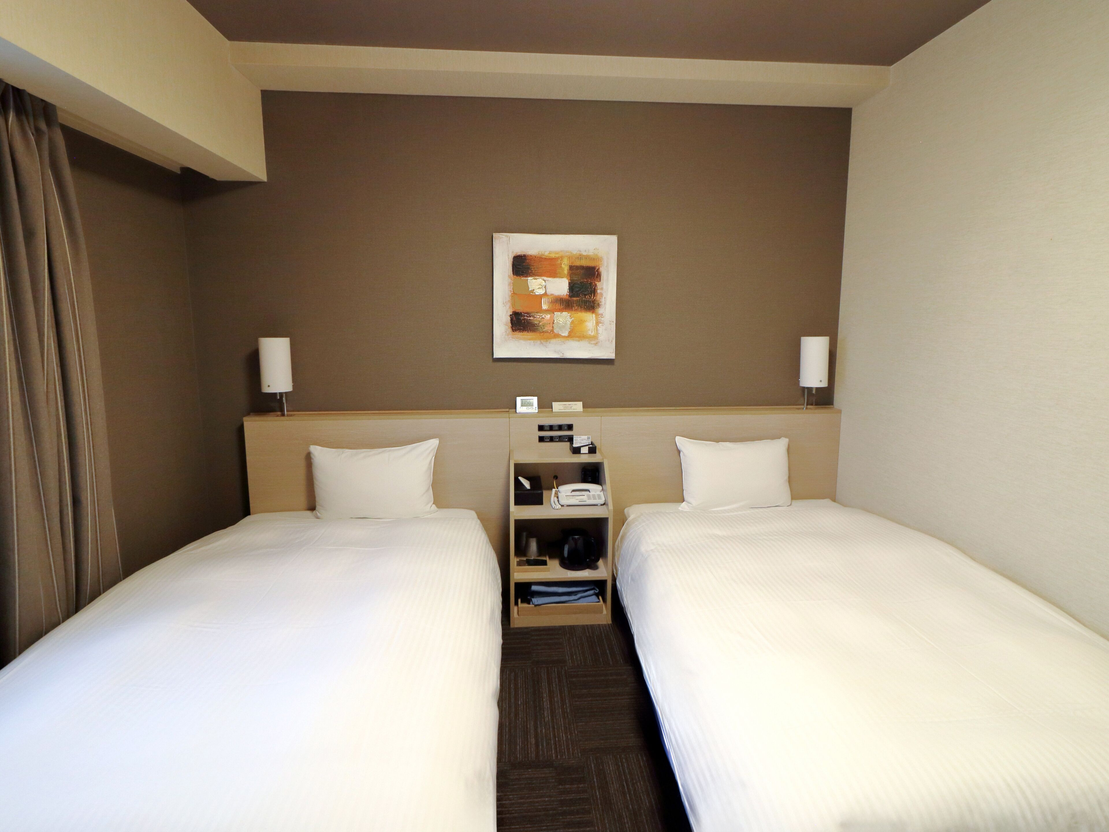 A hotel room with two single beds, a nightstand with a lamp, a small table with a phone and a clock, and a painting on the wall.