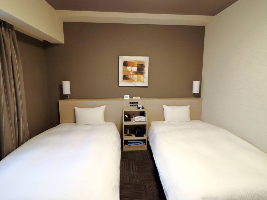 A hotel room with two single beds, a nightstand with a lamp, a small table with a phone and a clock, and a painting on the wall.