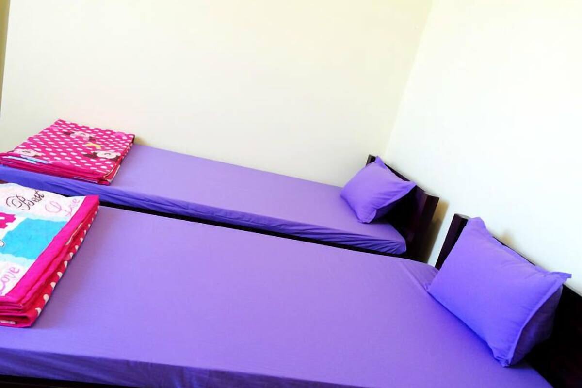 Zwei Einzelbetten mit violetter Bettwäsche und Kissen, eine pinkfarbene, gemusterte Decke und eine weiße Wand im Hintergrund.