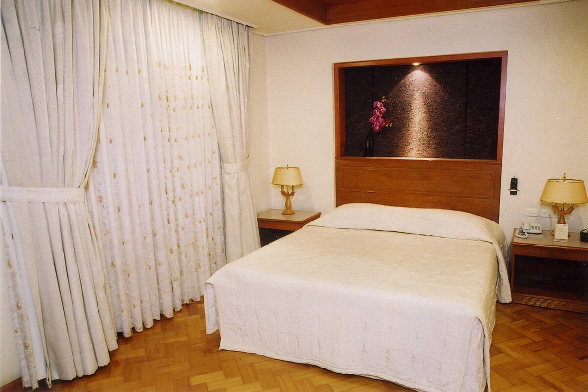 A hotel room with a large bed, a wooden headboard, two bedside tables with lamps, and a window with a floral decoration.