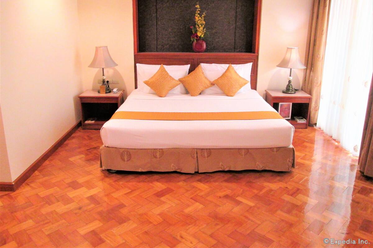A hotel room with a large bed, wooden floor, two bedside tables with lamps, a nightstand, and a window with curtains.