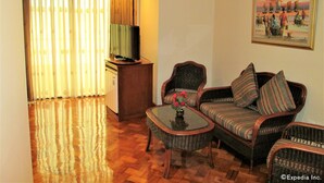 A living room with a television on a wooden stand, a sofa, two armchairs, a glass coffee table, and a painting on the wall. - Maxandrea Hotel (Cagayan de Oro)