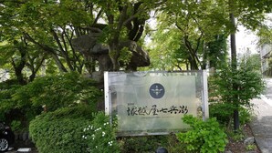 A sign with Japanese characters and a logo, surrounded by lush greenery and a stone pathway. - Tsukagoshiyashichibee (Shibukawa)