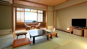 A traditional Japanese-style room with tatami flooring, a low table, cushioned seating, a television, and a view of the sea through shoji screens. - Tsukagoshiyashichibee (Shibukawa)