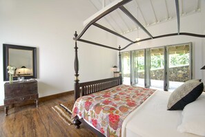 A four-poster bed with a floral bedspread, a wooden dresser with a mirror, and a view of a stone wall through open shutters. - Ivory House (Angulugaha)