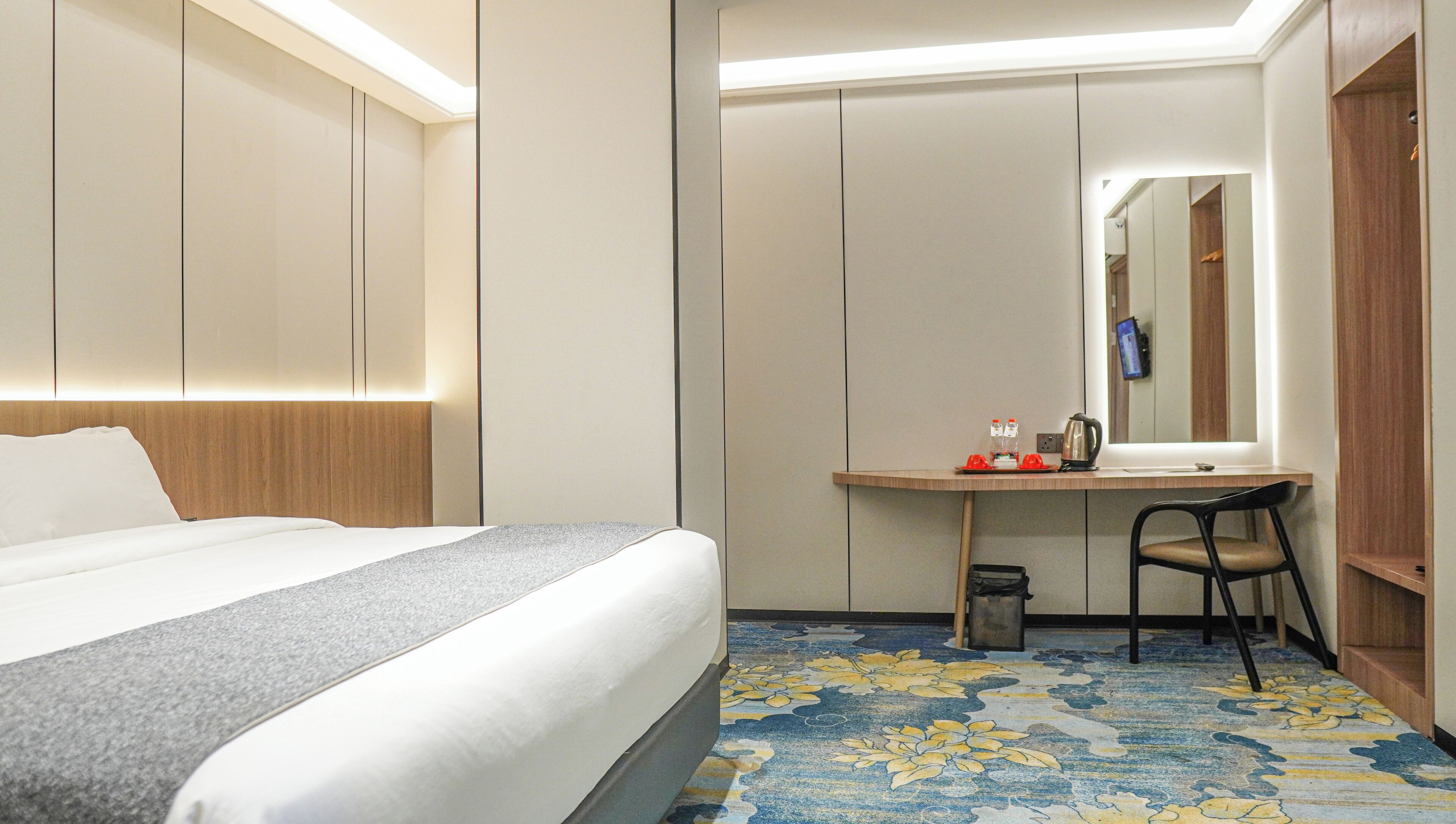 Una habitación de hotel con una cama, un escritorio con silla, un cesto de basura y un espejo.