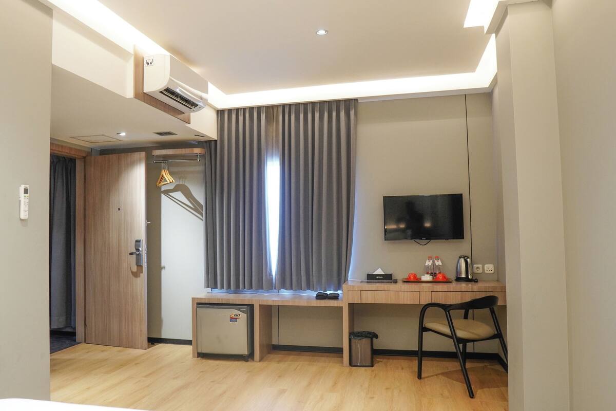 A hotel room with a wooden desk, a TV, a small fridge, a trash bin, and a chair.