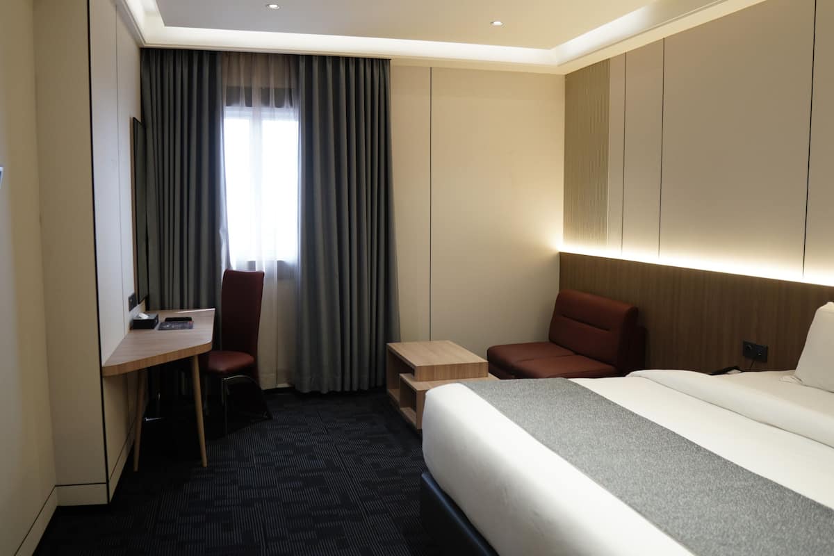 A hotel room with a bed, a desk, a chair, a TV, and a window with curtains.
