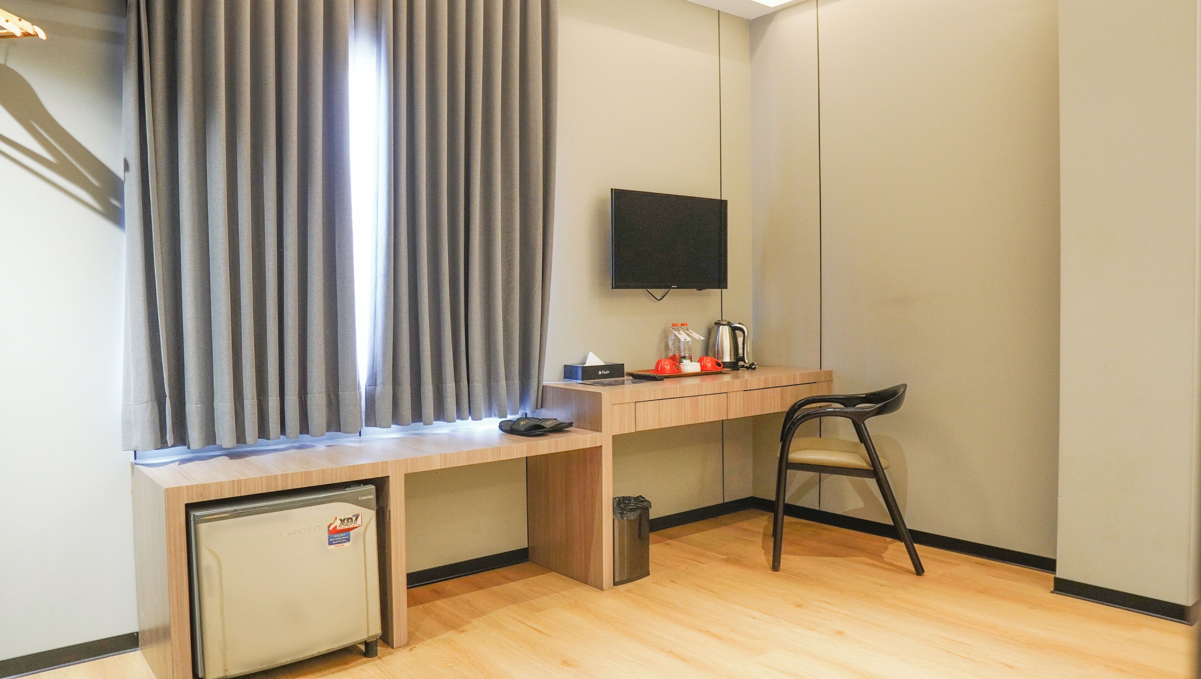 A compact hotel room with a wooden desk, a small refrigerator, a TV mounted on the wall, and a window with curtains.