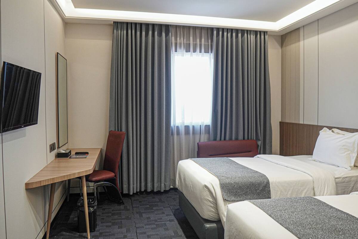 A hotel room with a desk, two beds, a TV, and a window with curtains.