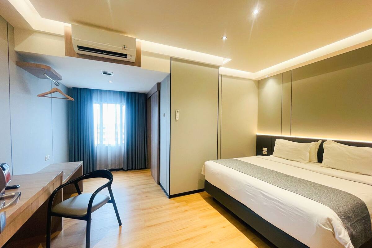 A modern hotel room with a bed, desk, chair, and an air conditioning unit.