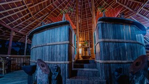 Une structure en bois surmontée d’un toit de chaume, comprenant deux grandes cuves cylindriques en bois, un escalier menant à un niveau supérieur et des sculptures en bois rustiques.
