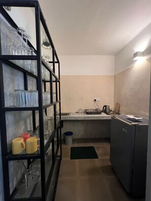 A compact kitchen with a built-in sink, a black metal shelf with glassware, a small refrigerator, and a countertop with various items. - Redcliffs by The Serendipity Collection (Mirissa)