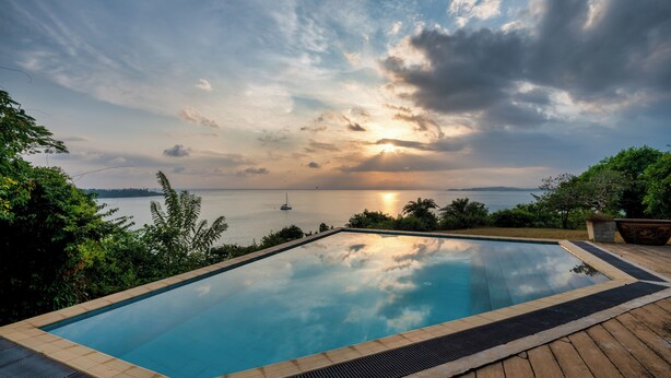 Ein rechteckiger Infinity-Pool mit Holzterrasse, umgeben von viel Grün, mit Blick auf ein Gewässer, auf dem ein Segelboot fährt, unter einer teilweise bewölkten Himmel.