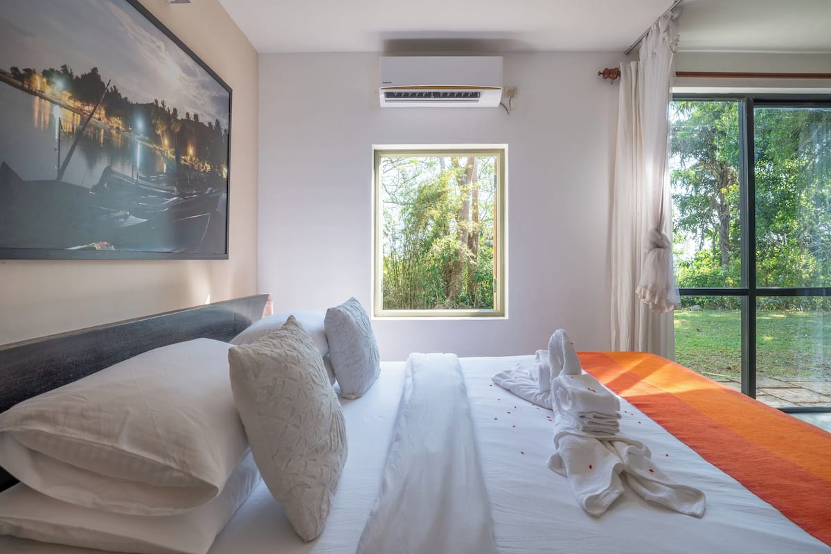 A hotel room with a large bed, a painting on the wall, a window with a view of greenery, and an air conditioning unit.
