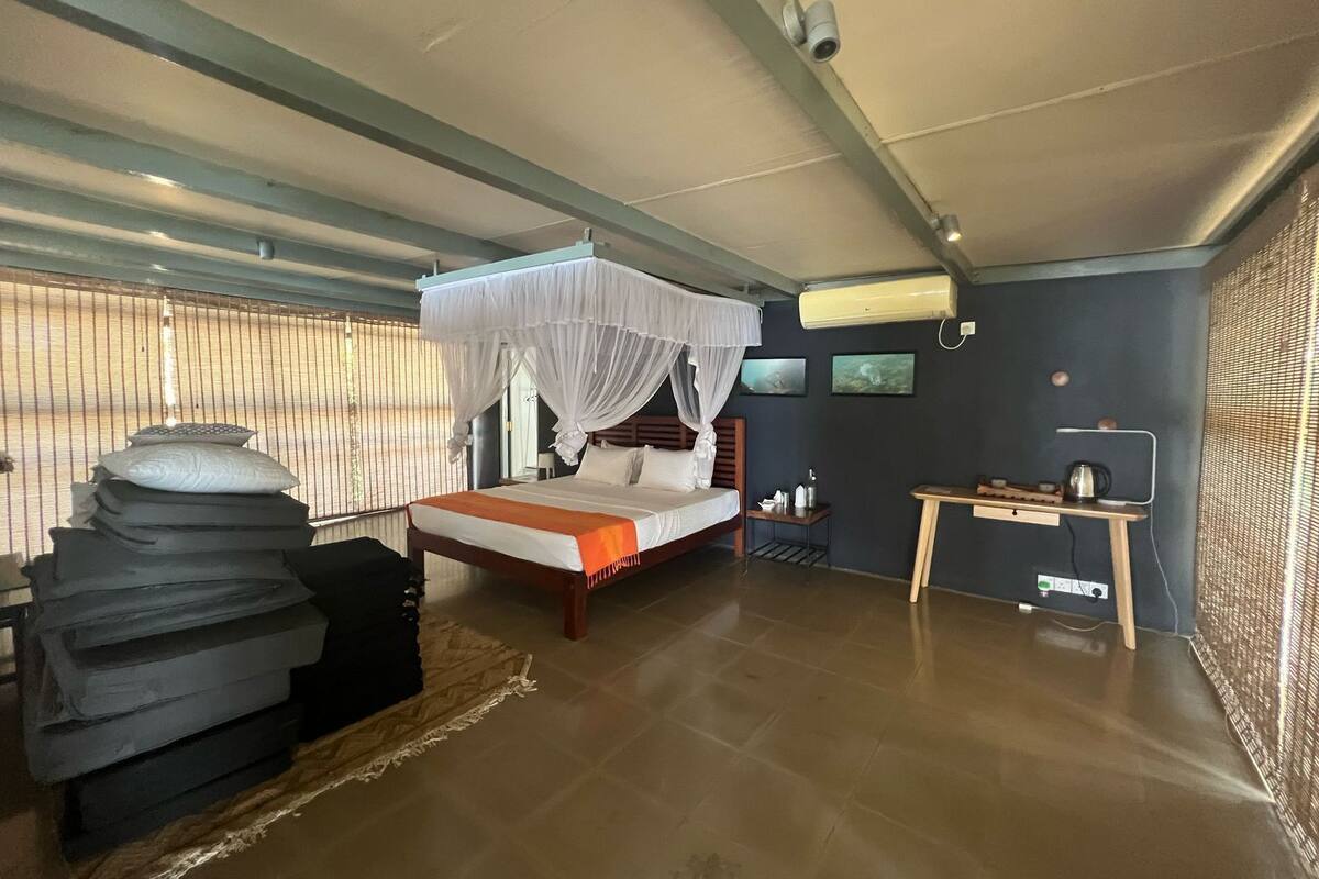A bedroom with a bed, a bedside table, a chair, a window with blinds, and an air conditioning unit.