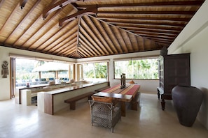 A spacious room with a high wooden ceiling, a large dining table, and a bench. There are windows providing natural light and a view of palm trees outside.