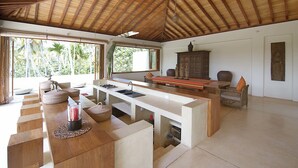 Una habitación amplia con techo de madera, una zona de cocina con lavaplatos y encimera, y grandes ventanales con vistas a palmeras.
