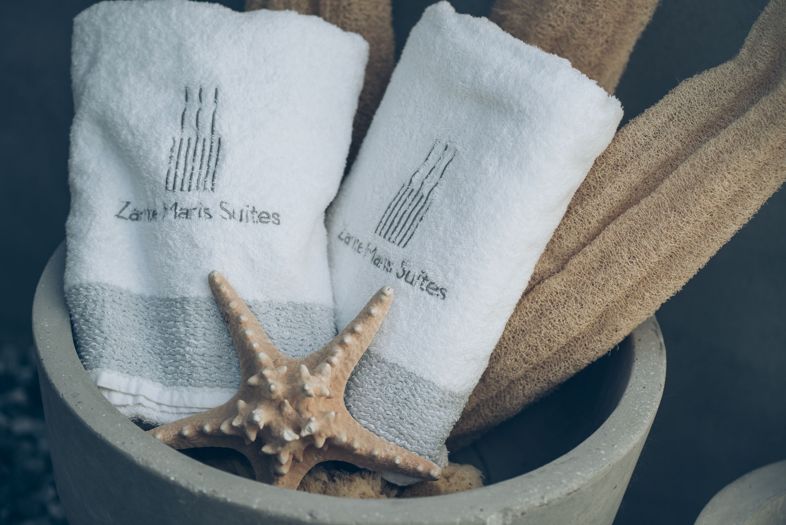 Two white towels with "Zane Maris Suites" embroidered on them, placed in a stone container with a starfish.