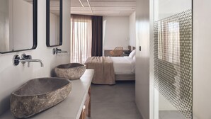 A modern hotel room with a large bed, a wooden bench, a stone basin, a large mirror, and a shower with a geometric pattern. - Zante Maris Suites - Adults Only (Zakynthos)