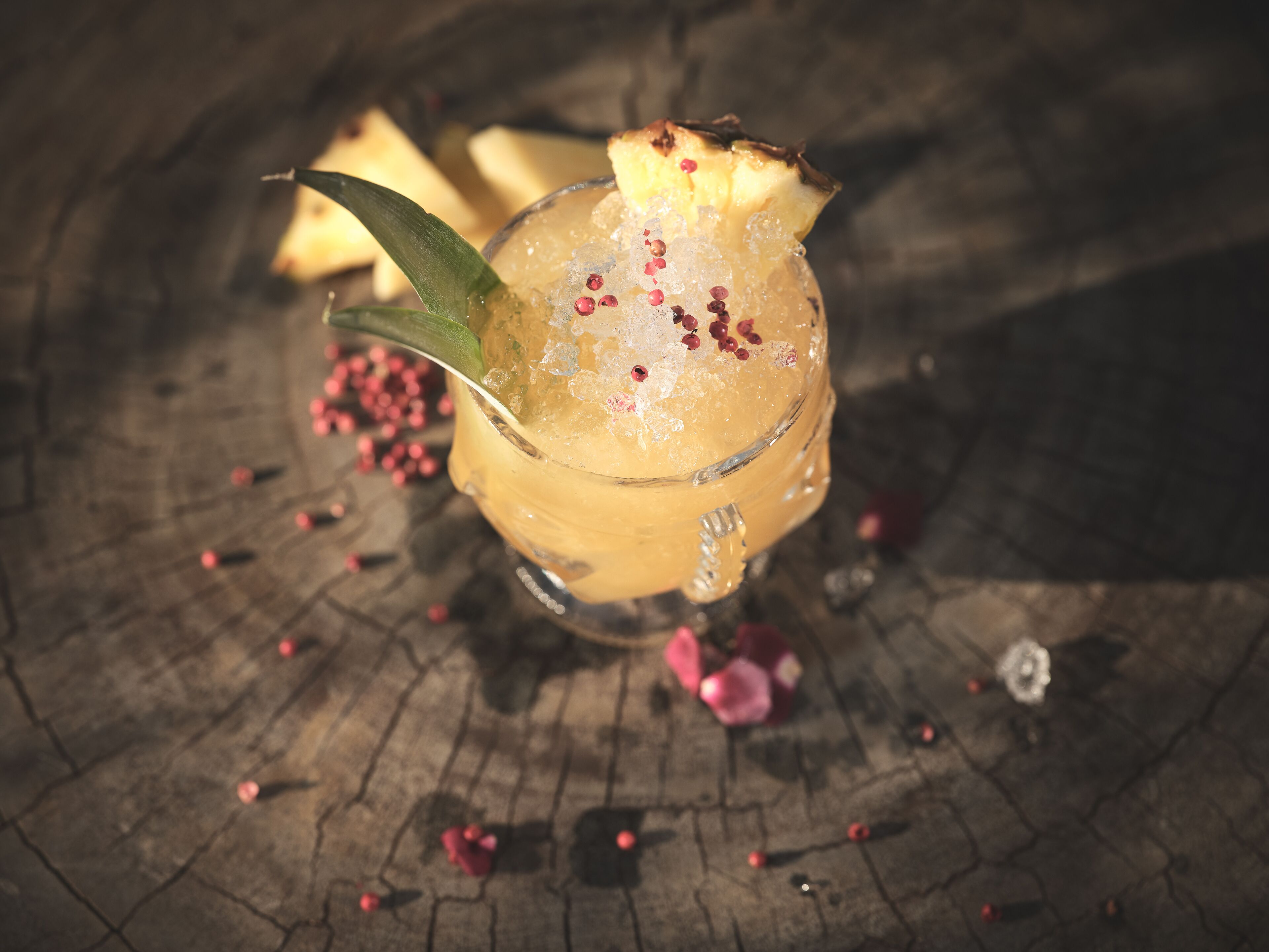 Un bicchiere con una fetta di ananas e un rametto di rosmarino, circondato da bacche rosse sparse su un tavolo di legno.