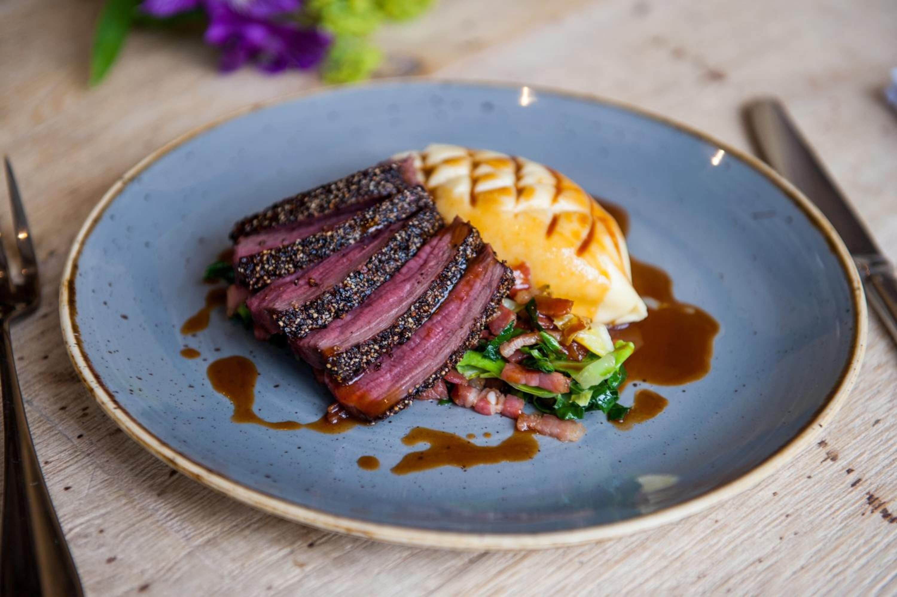 A plate with sliced meat, a grilled item, and greens on a blue plate.