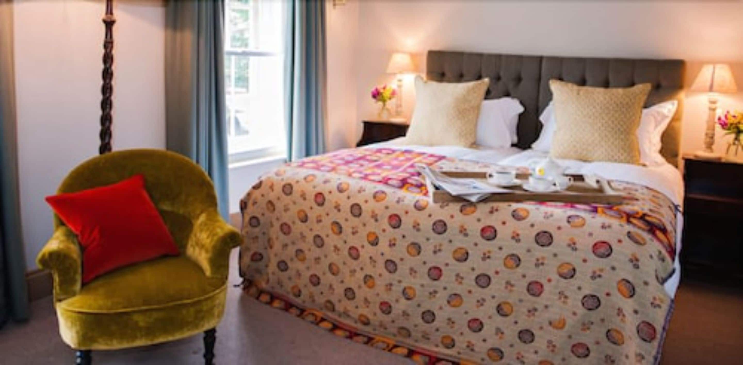 A hotel room with a large bed, a yellow armchair with a red pillow, and a window with curtains.