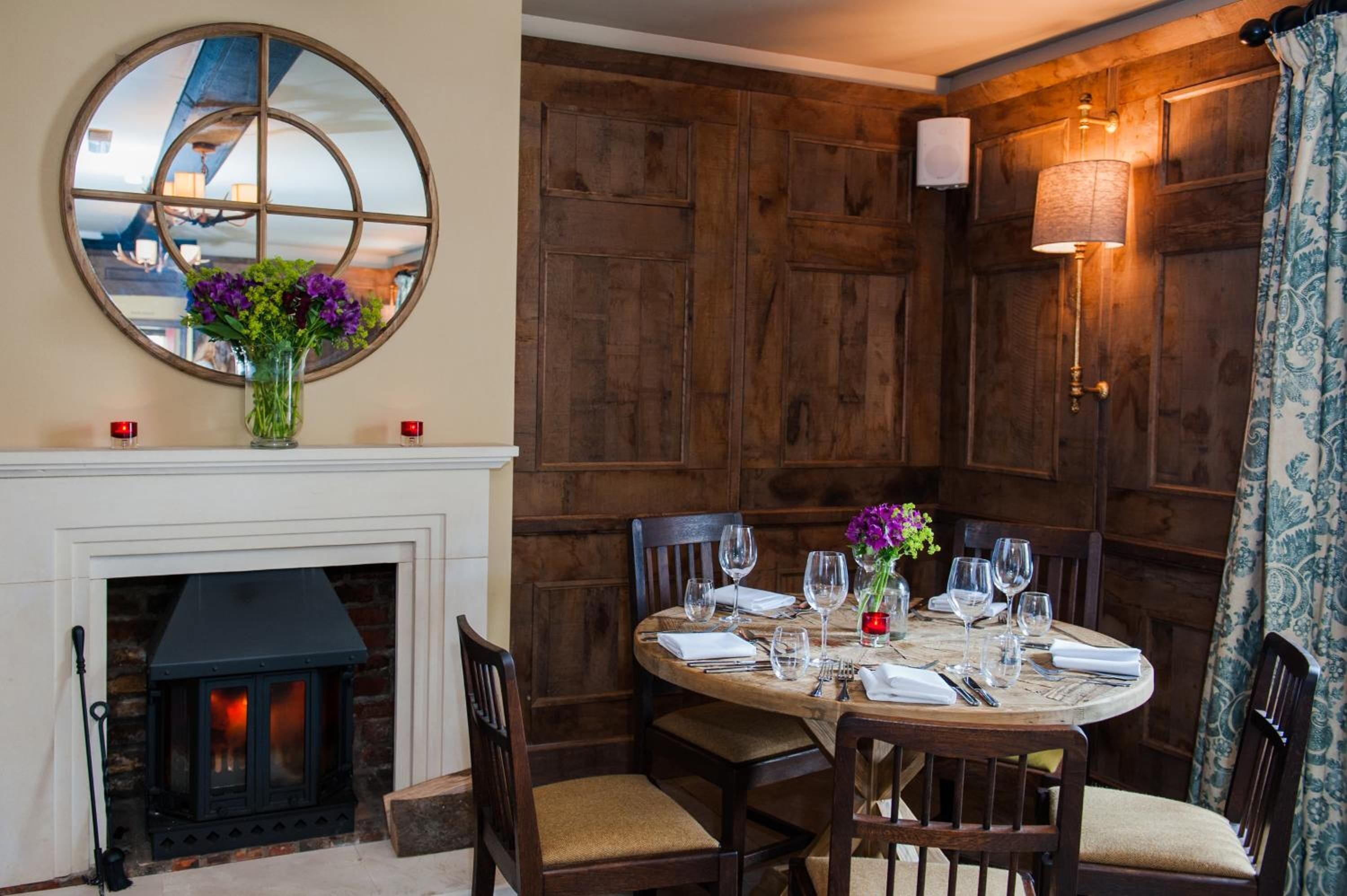 A dining area with a round wooden table set for four, surrounded by wooden chairs. A fireplace is visible to the left, and a mirror hangs above the table.