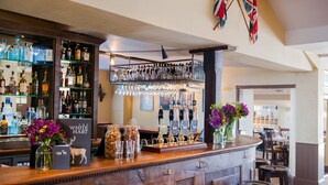 A bar area with a wooden counter, shelves with bottles, glasses, and a beer tap. There are flowers in vases and a sign reading "The White Hart".