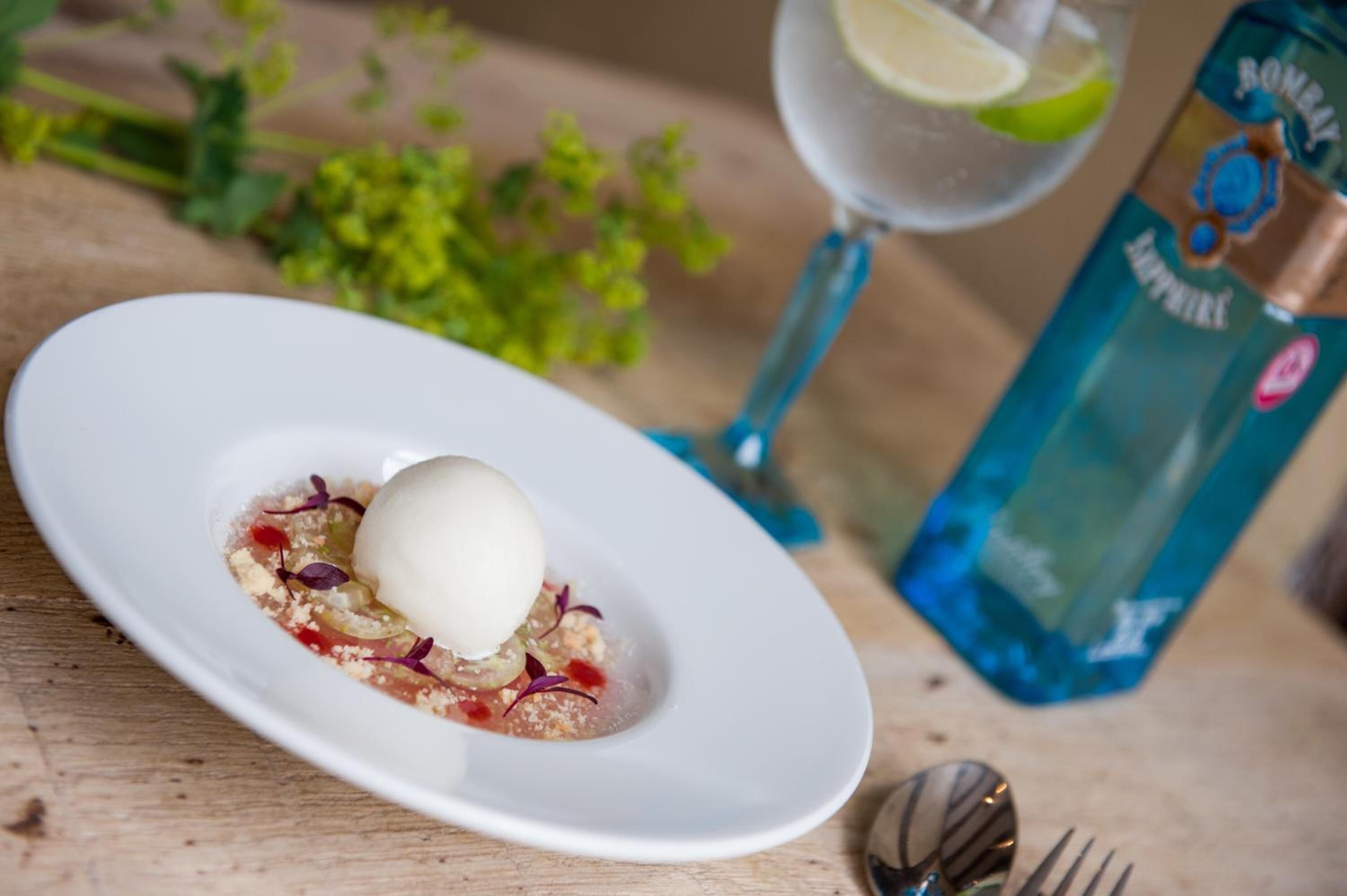 A white plate with a dessert, a glass of water with a lime slice, and a blue bottle on a wooden table.
