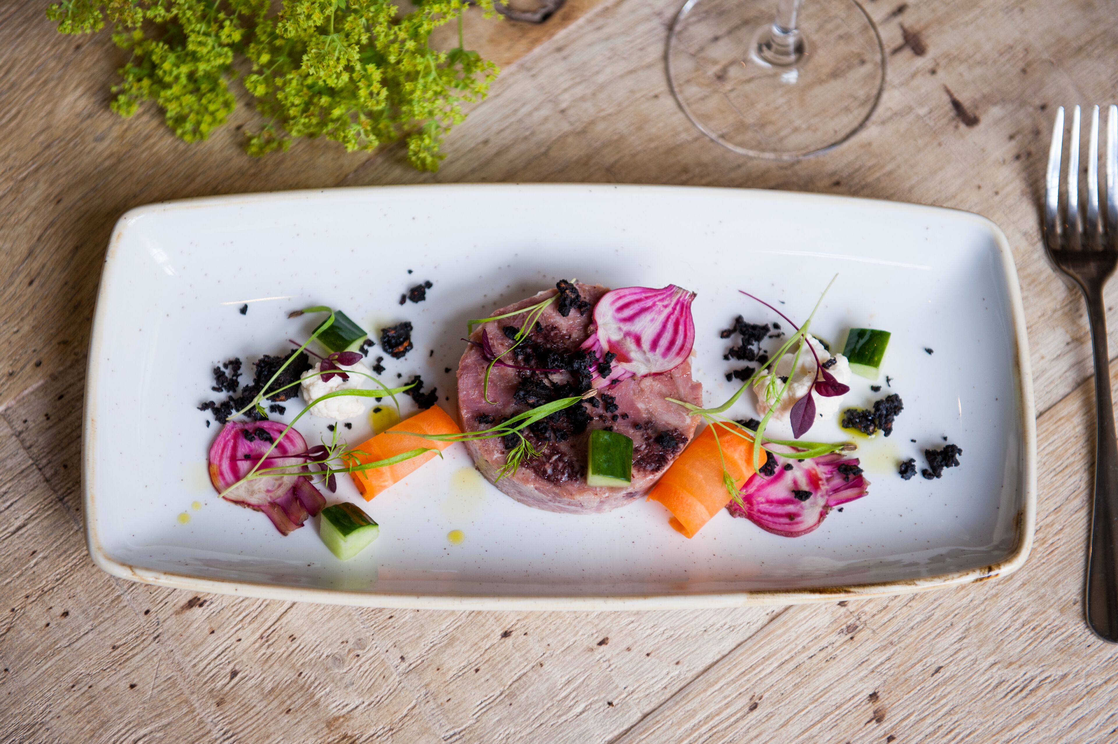 A rectangular white plate with a dish of meat, vegetables, and blackberries, accompanied by a fork and a glass of wine on a wooden table.
