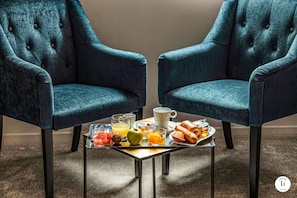 Two upholstered armchairs facing each other with a small table between them, holding breakfast items like toast, fruit, and beverages.
