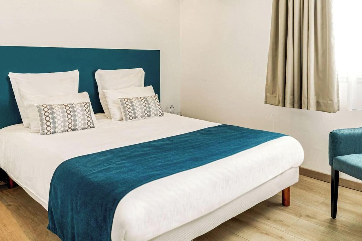 Una cama bien hecha con una manta azul, un sillón tapizado en color turquesa y una ventana con cortinas.