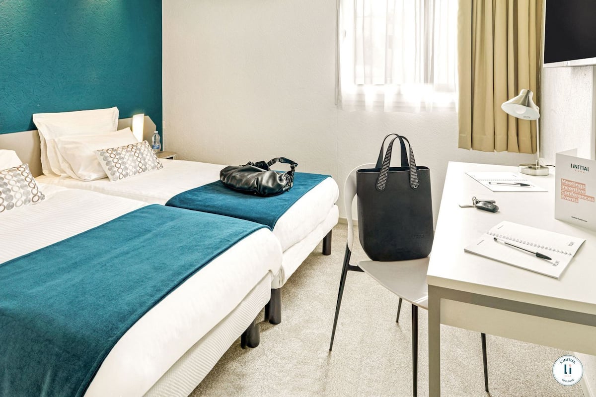 Habitación de hotel con dos camas, un escritorio con silla, una botella de agua, un bolso de mano y un cartel de L'Initial.